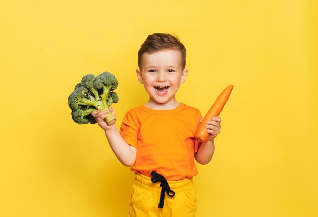 A smiling child holds a carrot and a head of broccoli