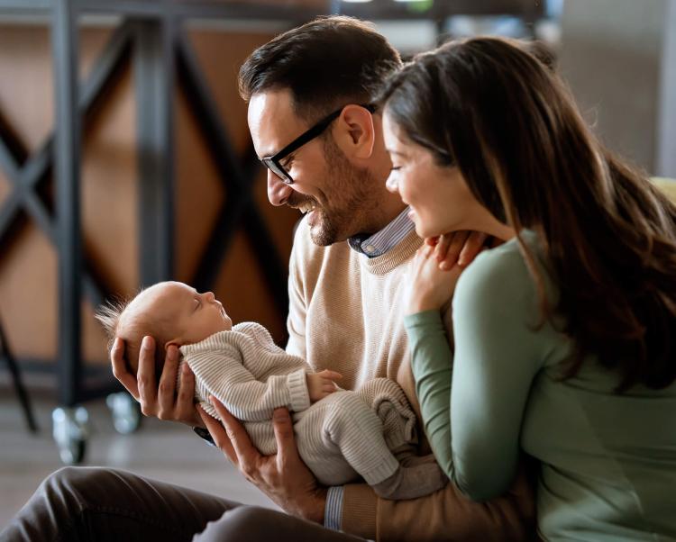 Mother and Father seated and staring down lovingly and smiling at their new baby being held in the father's arms. Kitchen island and stools visible in soft focus in background. 