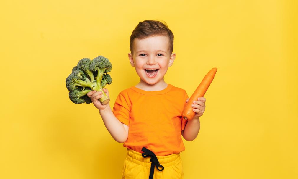 A smiling child holds a carrot and a head of broccoli