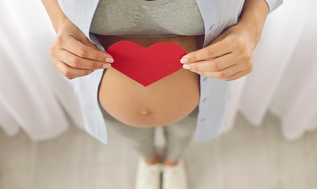 Closeup of a pregnant mother holding a red heart-shaped card over her baby bump
