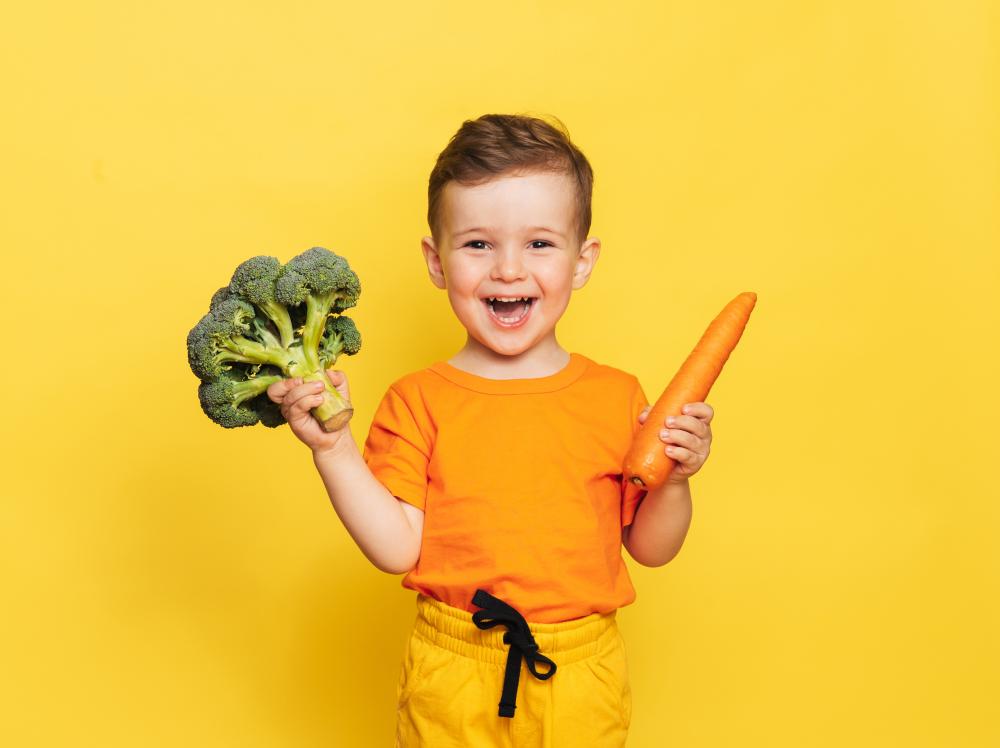 A smiling child holds a carrot and a head of broccoli