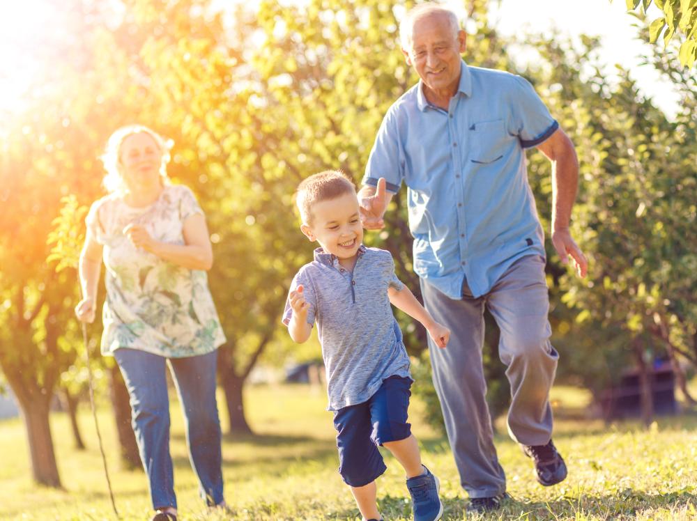 mature couple in sunny meadow chases laughing grandson